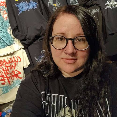 Katri is smiling while standing in front of a various assortment of hanging band t-shirts.