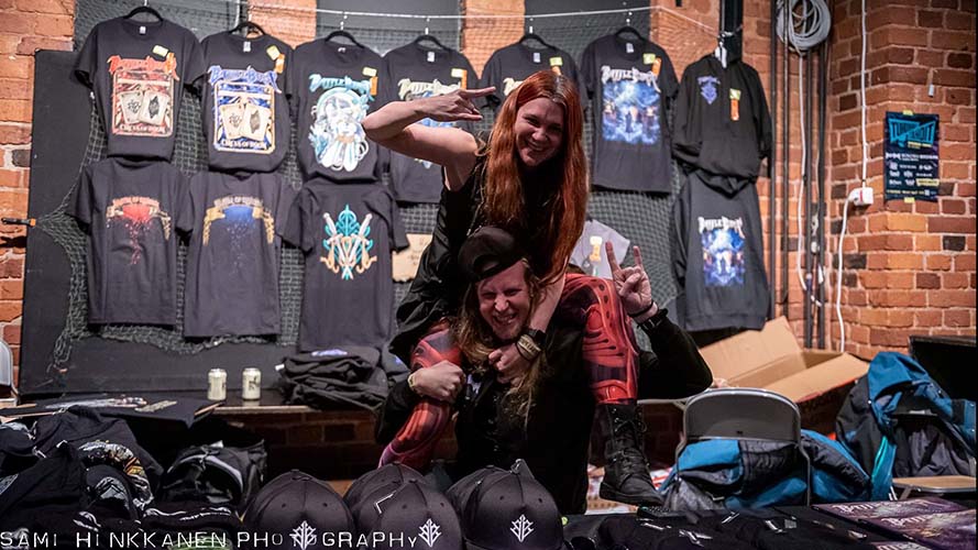 Chrissy is sitting on Miikka's shoulders in front of a wall full of hanging Battle Beast shirts. They're both having fun and laughing.