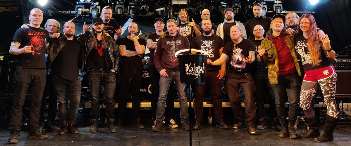 The bands Kalmah, Mors Subita, and Suotana, plus their support crew are standing on the stage at Jyväskylä's Tanssisali Lutakko venue. A black bucket with a Kalmah logo is hanging on a microphone stand, front and center.