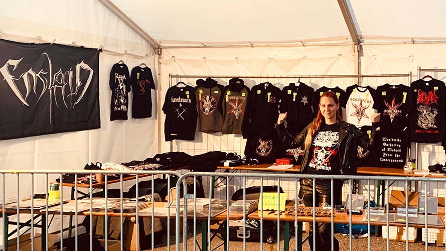Chrissy Hujanen standing in a tent at a metal festival behind a table full of band merchandise.