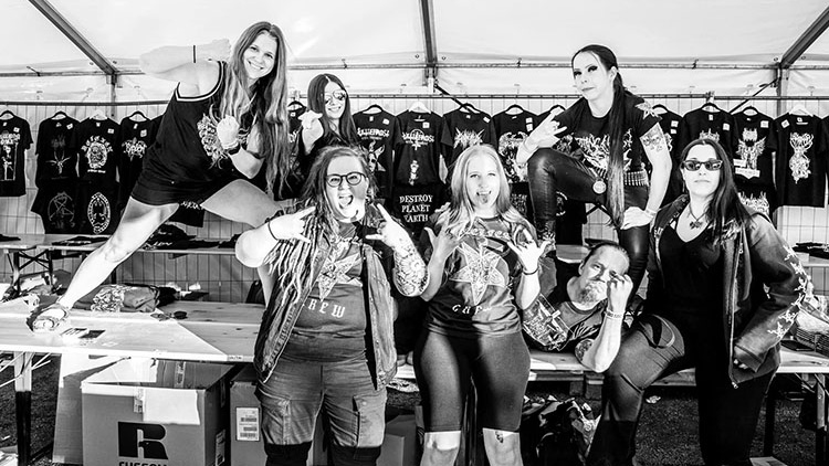 Chrissy, Nadya, Katri, Milla, Serena, Janetta and Aaro are huddled together and smiling, standing in front of and top of the merchandise table at Steelfest 2023.