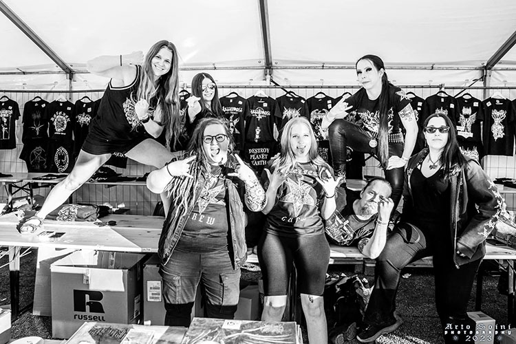 Chrissy, Nadya, Janetta, Katri, Milla, Aaro and Serena are huddled together and smiling, standing in front of and top of the merchandise table at Steelfest 2023.