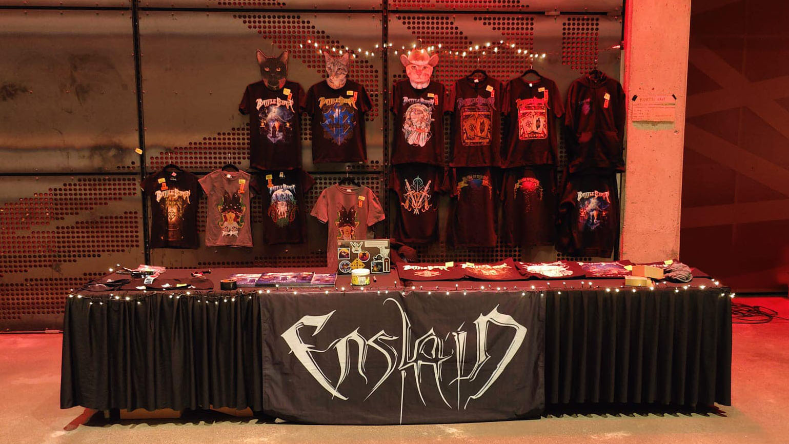 Large display of Battle Beast merchandise consisting of t-shirts and hoodies hanging on a wall, as well as vinyls, fabric shopping bags and beanines on a tablet. Enslain's company banner is hanging off the side of the table.